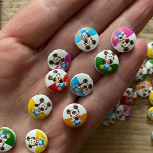 May include: A selection of small, round wooden buttons featuring Mickey Mouse. Each button has a white background with a colourful border in shades of blue, pink, yellow, orange, and green. The buttons are approximately 2 cm in diameter.