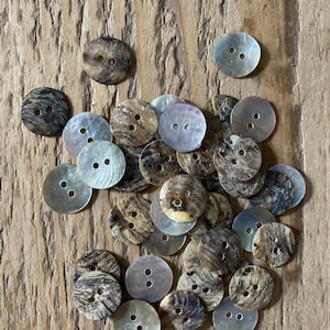 Puede incluir: Un conjunto de botones redondos, cada uno con dos agujeros, en tonos naturales variados: marrón, beige y nacarado. Los botones están dispuestos sobre una superficie de madera, resaltando sus texturas y patrones únicos, ideales para costura.