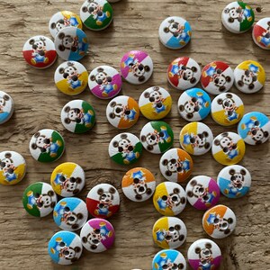 Può includere: Una collezione di bottoni in legno con un design a personaggio dei cartoni animati. Ogni bottone presenta uno sfondo bianco con un bordo segmentato colorato in vari colori e un personaggio dei cartoni animati al centro. I bottoni sono sparsi su una superficie di legno.
