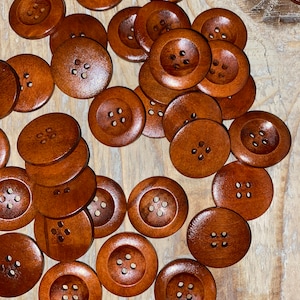 Może przedstawiać: Kolekcja okrągłych, drewnianych guzików w różnych rozmiarach. Każdy guzik ma cztery otwory do szycia i bogaty, brązowy kolor. Guziki są rozrzucone na jasnej drewnianej powierzchni, ukazując naturalne usłojenie drewna.