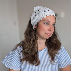 May include: A white lace headscarf featuring an intricate floral pattern and a scalloped edge, worn tied over long wavy brown hair. The headscarf is paired with a light blue and white vertically striped short-sleeved top with ruffled sleeves.