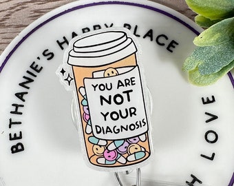 May include: White round badge holder with a purple border. The badge holder has the text "Bethanie's Happy Place Made With Love" around the edge. A clear plastic badge holder with a cartoon image of a coffee cup with pills inside and the text "You are not your diagnosis" is attached to the badge holder.