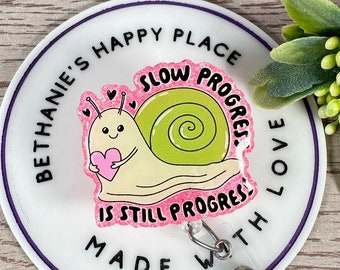 Könnte beinhalten: Ein runder weißer Badgehalter mit einem violetten Rand. Er zeigt eine rosa und grüne Schnecke mit einem Herzen und dem Text "Bethanie's Happy Place Slow Progress Is Still Progress" und "Made With Love".