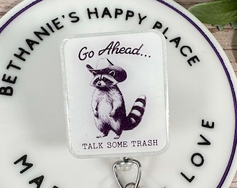 May include: A white and purple badge reel with the text "BETHANIE'S HAPPY PLACE MADE WITH LOVE". The badge reel has a rectangular design with a raccoon wearing a cowboy hat and the text "Go Ahead... TALK SOME TRASH".