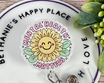 Könnte beinhalten: Ein runder weißer Badgehalter mit einem violetten Rand und dem Text "Bethanie's Happy Place" und "Made With Love". Der Badgehalter enthält einen durchsichtigen Plastikbadge mit einer gelben Sonnenblume mit einem lächelnden Gesicht und dem Text "Mental Health Matters".