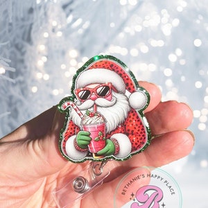 Könnte beinhalten: Ein roter und grüner Glitzer-Weihnachtsmann mit Sonnenbrille hält einen roten und weißen Milchshake. Der Weihnachtsmann trägt einen roten und weißen Hut und einen roten und weißen Anzug mit grünem Glitzerbesatz. Der Weihnachtsmann hält einen roten und weißen Strohhalm im Mund. Der Weihnachtsmann ist an einem durchsichtigen, ausziehbaren Badgehalter aus Plastik befestigt, auf dem der Text "Bethanie's Happy Place Made With Love" in einem rosa Kreis mit einem rosa Buchstaben B in der Mitte steht.