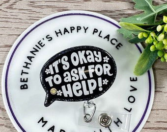 May include: White round badge holder with purple trim and the text "Bethanie's Happy Place Made With Love" around the edge. A black and glitter speech bubble with the text "It's okay to ask for help!" is attached to the badge holder.
