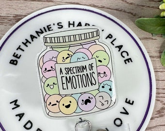 May include: A white round badge holder with a purple border. The badge holder has the text "Bethanie's Happy Place Made With Love" around the edge. In the center is a clear plastic badge holder with a cartoon image of a jar filled with colorful cartoon faces. The text "A Spectrum of Emotions" is on the jar.