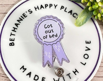 Könnte beinhalten: Ein runder weißer Badgehalter mit violetter Umrandung und dem Text "Bethanie's Happy Place Made With Love". Ein violettes Band mit Glitzer und dem Text "Got out of bed" ist am Badgehalter befestigt.