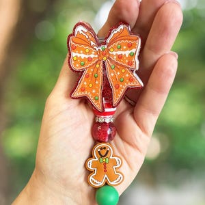 Könnte beinhalten: Ein festliches Ausweis-Rollband mit Lebkuchenmann und Schleifendesign. Die Schleife ist orange mit roten Glitzerakzenten und bunten Punktdetails. Der Lebkuchenmann ist braun mit einer grünen Fliege. Eine grüne Perle und ein Schlüsselring vervollständigen das Design.