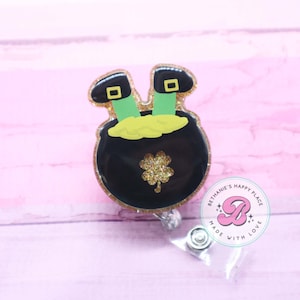 May include: A black pot of gold with a green shamrock and two green leprechaun legs sticking out of the top. The pot is decorated with gold glitter. The image also includes a round sticker with the text "Bethanie's Happy Place" and a stylized letter "B".