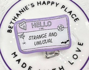 May include: A white round badge with a purple border that says "Bethanie's Happy Place Made With Love".  The badge has a purple and white ID card attached to it that says "Hello I am... Strange and Unusual". The ID card has a black and white Ouija board heart, a black pentagram, and a black bat.