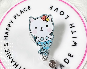 May include: White and blue mermaid cat badge holder with glitter. The badge holder has a clear plastic backing and a metal clip. The text on the badge holder reads "Bethanie's Happy Place Made With Love".