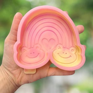 Könnte beinhalten: Eine rosa und gelbe Silikonform mit einem Regenbogen-Design, zwei lächelnden Froschgesichtern an der Basis und einem kleinen Herz in der Mitte. Die Form hat eine glatte, flexible Textur und einen erhöhten Rand zum Gießen von Formen.