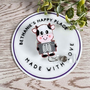May include: A white and black cow cartoon with pink accents holding a gray and white x-ray image. The cow is on a white round badge with the text "Bethanie's Happy Place Made With Love" in black text with a purple outline.