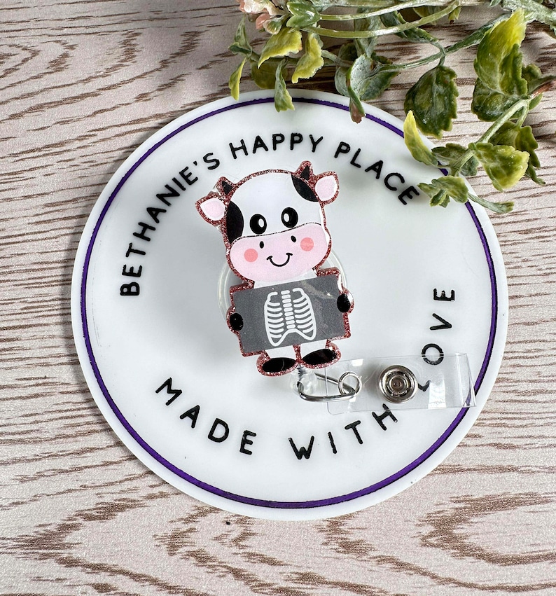 May include: A white and black cow cartoon with pink accents holding a gray and white x-ray image. The cow is on a white round badge with the text "Bethanie's Happy Place Made With Love" in black text with a purple outline.
