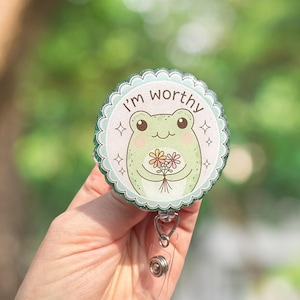 May include: A round badge reel with a cute green frog holding flowers. The badge reel has the text "I'm worthy" and a scalloped edge. The badge reel is attached to a clear plastic holder with a metal clip.