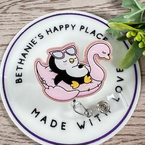 May include: A round white badge with a purple border featuring a cartoon penguin wearing sunglasses, floating on a pink swan float. The text "BETHANIE'S HAPPY PLACE" and "MADE WITH LOVE" are printed on the badge.
