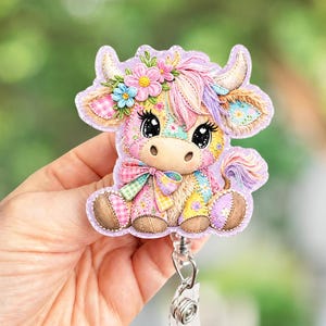 May include: A colorful cow badge reel with a patchwork design. The cow has large eyes, a floral crown, and a bow. The badge reel is attached to a retractable cord and a clear plastic holder.