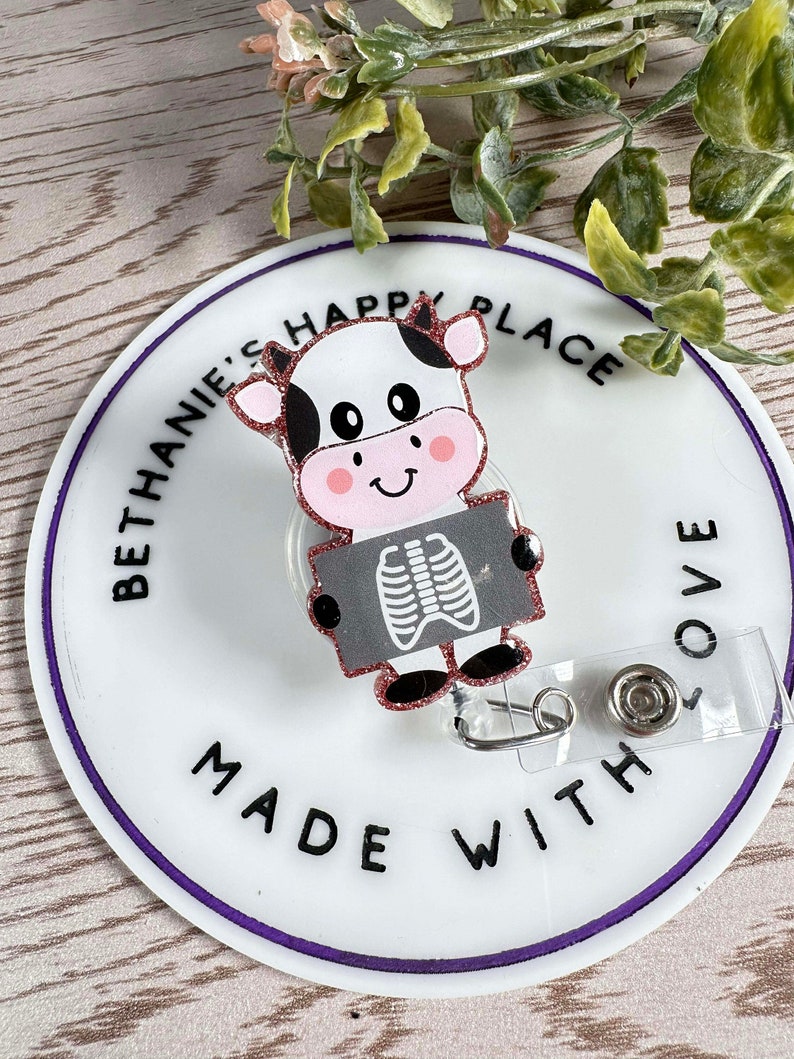 May include: A white and black cow cartoon with pink cheeks and a black and white x-ray image. The cow is holding the x-ray image. The background is a white circle with the text "Bethanie's Happy Place Made With Love" in black.