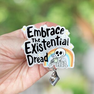 May include: A badge reel with the text "Embrace The Existential Dread" in black lettering. The badge features a skeleton drinking from a mug, with a rainbow behind it. The badge has a silver glitter background and a clear plastic coating.