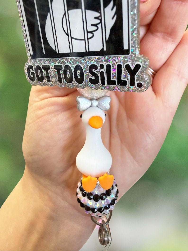 K&ouml;nnte beinhalten: Ein wei&szlig;es, g&auml;nsef&ouml;rmiges Ausweis-Jojo mit orangefarbenen F&uuml;&szlig;en und einer kleinen Schleife. Das Ausweis-Jojo hat den Text "GOT TOO SILLY" auf einem schwarz-wei&szlig;en Hintergrund. Das Jojo ist mit Glitzer verziert.