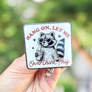 May include: A white square badge with a black and white illustration of a raccoon wearing heart-shaped sunglasses and holding a boba tea. The text on the badge reads "Hang on, let me overthink this".