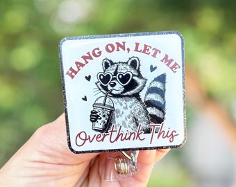 May include: A white square badge with a black and white illustration of a raccoon wearing heart-shaped sunglasses and holding a boba tea. The text on the badge reads "Hang on, let me overthink this".