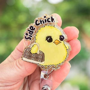 May include: A gold glitter badge holder with a cartoon yellow chick holding a brown and gold purse. The chick has large black eyes and eyelashes. The text "Side Chick" is written in black letters on the badge holder.