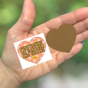 May include: Two heart-shaped stickers. One is brown, the other is white with an orange and pink checkered design. The white sticker has the text "FUNCTIONING ON SARCASM AND SNACKS" and a smiling face.