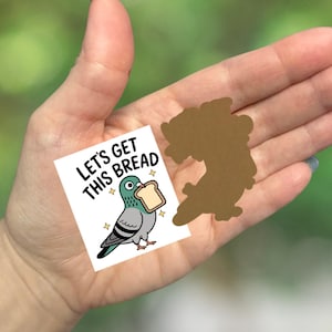 May include: A white sticker with the text "LET'S GET THIS BREAD" and a cartoon pigeon holding a slice of bread. A brown silhouette of a shape is next to the sticker. The background is a blurred green.