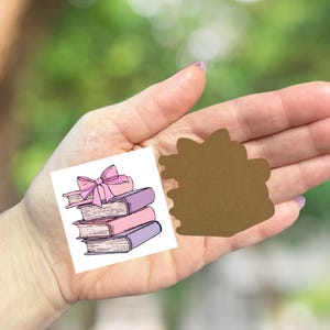 May include: A small, square illustration of a stack of books with a pink bow on top. The books have pink, purple, and brown covers. A brown, die-cut shape is next to the illustration. The background is a blurred green.