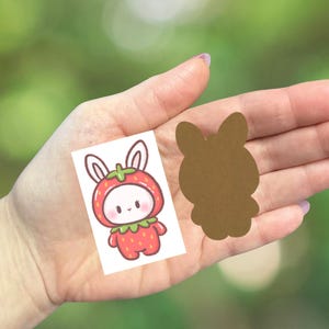 May include: A small, rectangular sticker featuring a cute cartoon bunny wearing a strawberry costume. Next to it is a brown bunny-shaped cutout. The background is a blurred green.