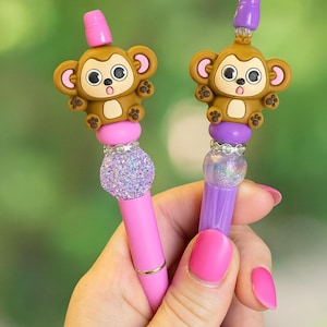May include: Two decorative pens with monkey toppers. One pen is pink with a glittery silver accent, and the other is purple with a clear, glittery accent. Both pens have a brown monkey face with large eyes and a pink top.