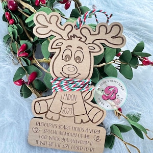 May include: A wooden reindeer ornament with a red and white striped scarf. The ornament is personalized with the name Landon and the year 2023. The ornament has a message that reads "Rudolph's scarf holds a very special memory of me. For when you unwrap it, it will be the size I used to be."