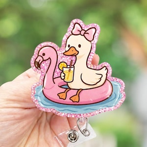 May include: A whimsical badge reel featuring a cartoon duck with a pink bow, floating on a pink flamingo pool float. The duck holds a yellow drink with a lemon slice. The design has a glittery pink border and a blue water background.