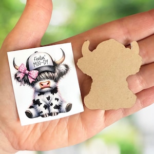 May include: A printed sticker and a wooden cutout of a Highland cow. The sticker features a cartoon cow wearing a hat with the text "Feelin' Moo-dy" and a pink bow. The cow has black and white spots and long horns. The wooden cutout is in the shape of a cow.