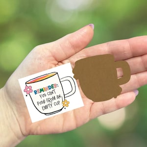 May include: A white mug illustration with a rainbow-colored interior and the text "REMINDER: You can't pour from an empty cup." A brown mug cutout is also present. The background is a blurred green.