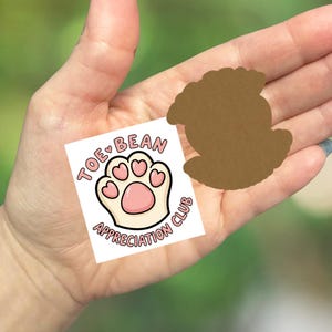 May include: A white sticker with a paw print design and the text "Toe Bean Appreciation Club" in pink and black. A brown die-cut shape of a dog's paw is next to the sticker.