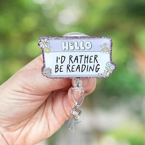 May include: A light purple retractable badge reel with the text "HELLO I'D RATHER BE READING". The badge reel has a glittery border with flower and rainbow illustrations. The reel is held by a hand against a blurred green background.