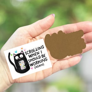 May include: A white sticker with a black cat illustration holding a phone, with the text "SCROLLING WHEN I SHOULD BE WORKING (AGAIN)". A brown die-cut shape is also visible.