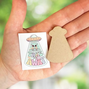 May include: A small, rectangular sticker with a colorful illustration of a UFO and an alien, with the text "TAKE ME AWAY". Beside it is a small, unfinished, UFO-shaped wooden cutout. The items are held in a hand.