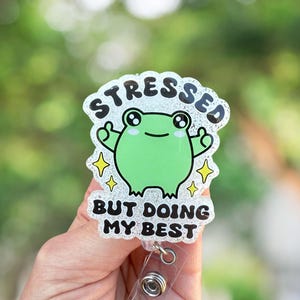 May include: A green frog cartoon character with glitter on a white background. The frog is giving a thumbs up with both hands. The text reads "Stressed but doing my best". The image shows a badge holder with a clear plastic strap and a metal clip.