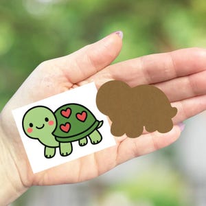 May include: A green cartoon turtle with a smiling face and red heart details on its shell. Next to it is a brown turtle-shaped cutout. The image is held in a hand.