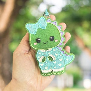 May include: A glittery green dinosaur-shaped keychain with a light blue bow and matching pajamas. The dinosaur has a friendly face with black eyes and a smile. The pajamas are decorated with white hearts.