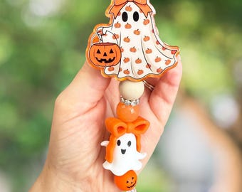 May include: A Halloween-themed badge reel featuring a ghost design. The ghost is white with orange pumpkin accents and a bow. The reel also includes a smaller ghost, pumpkin, and decorative beads. The reel is attached to a silver clip.