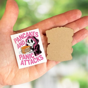 May include: A white sticker with the text "PANCAKES AND PANIC ATTACKS" and a cartoon grim reaper character holding pancakes. Next to it is a blank, brown, irregularly shaped wooden cutout.