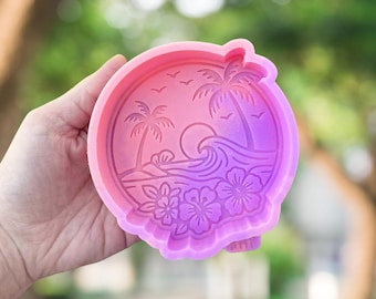 May include: A pink and purple silicone mould featuring a tropical beach scene. The design includes palm trees, waves, flowers, and birds. The mould is round with a scalloped edge.