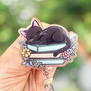 May include: A black cat sleeping on top of a stack of books with floral accents. The books are blue, pink, and beige. The cat has pink ears and a pink nose. The image is on a clear plastic badge holder with a retractable reel.
