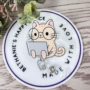 May include: White round badge holder with a purple border. The badge holder features a cartoon cat wearing glasses and using a laptop. The text "BE HAPPY" is on the left side of the badge holder and "WITH LOVE" is on the right side. The text "MADE" is on the bottom of the badge holder.